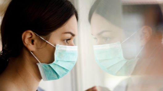 portrait-of-sad-young-woman-with-face-protective-mask-looking-through-window