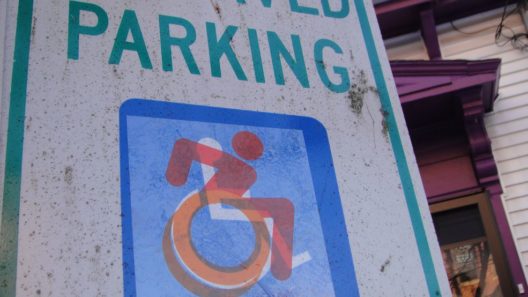original International Symbol of Access on a 'reserved parking' sign, with a red and orange, leaning forward, “italicized,” sticker by the Accessible Icon Project overlaid