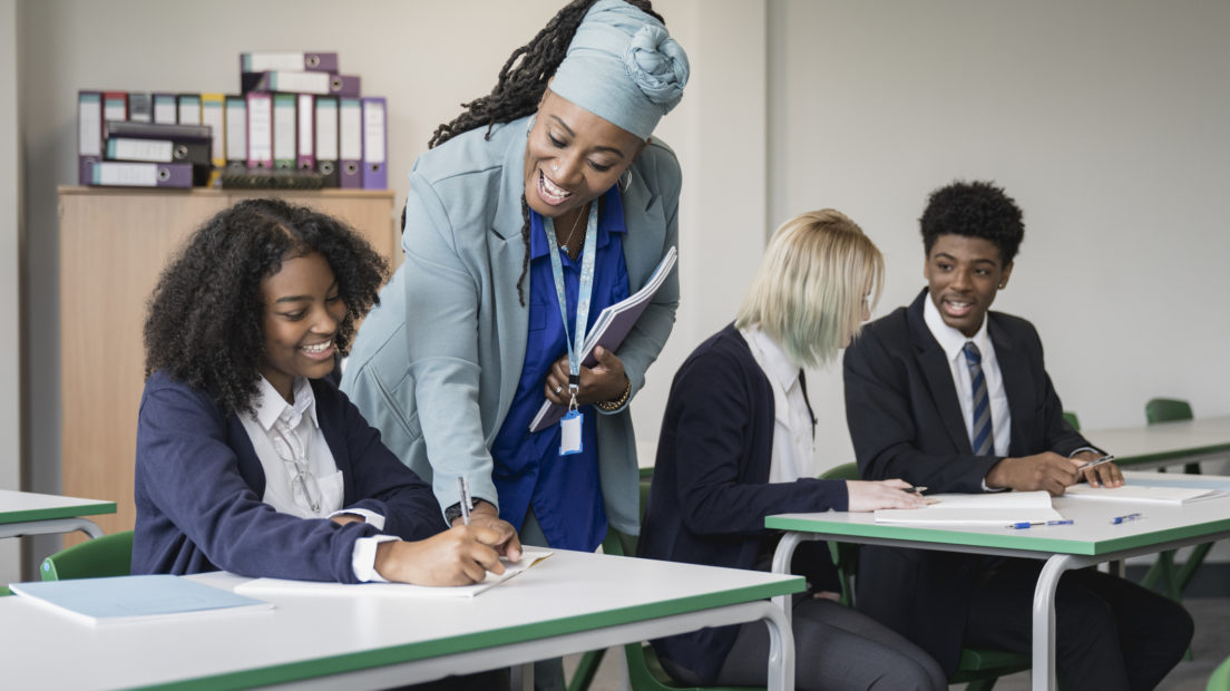 3 teenage students wearing school uniforms sat at desks. Two are chatting while one speaks to their teacher.