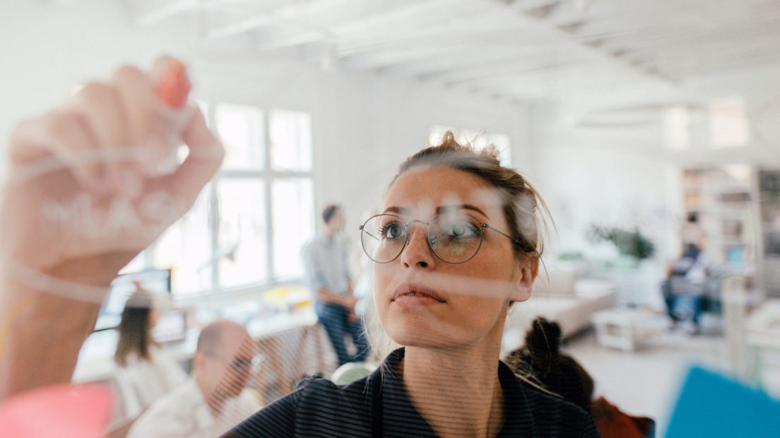 Photo of a young woman writing on a transparent wipe board and thinking of a solution for her work-related problems