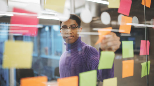 A man in a purple jumper looks at different coloured post-it notes that are on a glass wall