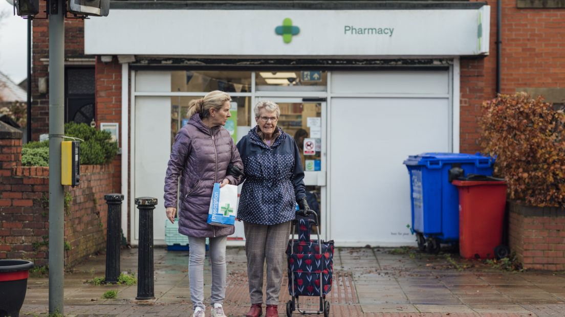 An elderly woman linking arms with her daughter, stood outside a pharmacy in the UK