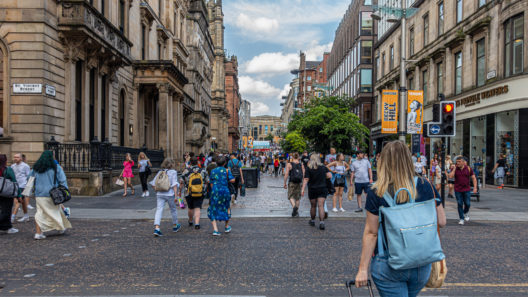 People walking along the busy pedestrianised shopping street, Buchanan Street, in the centre of Glasgow, Scotland. At the far end of the road is the Royal Concert Hall, behind an entrance to Buchanan Street Underground Station.