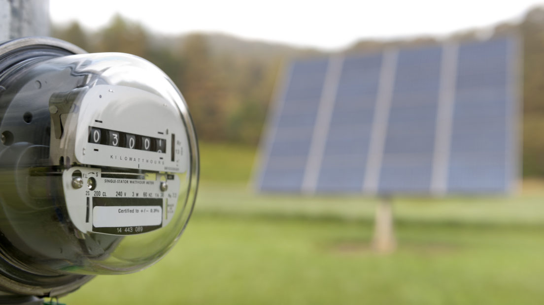 An electric meter with an out of focus solar panel behind it.