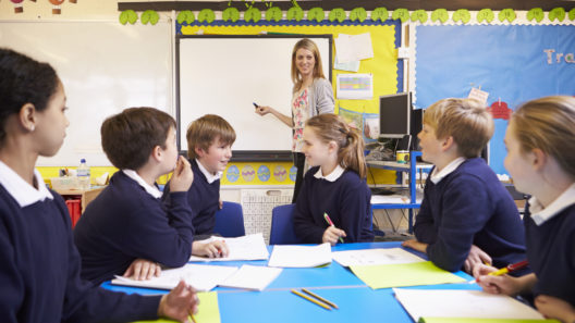Pupils Sitting At Table As Teacher Stands By Whiteboard Writing Up Lesson Objectives