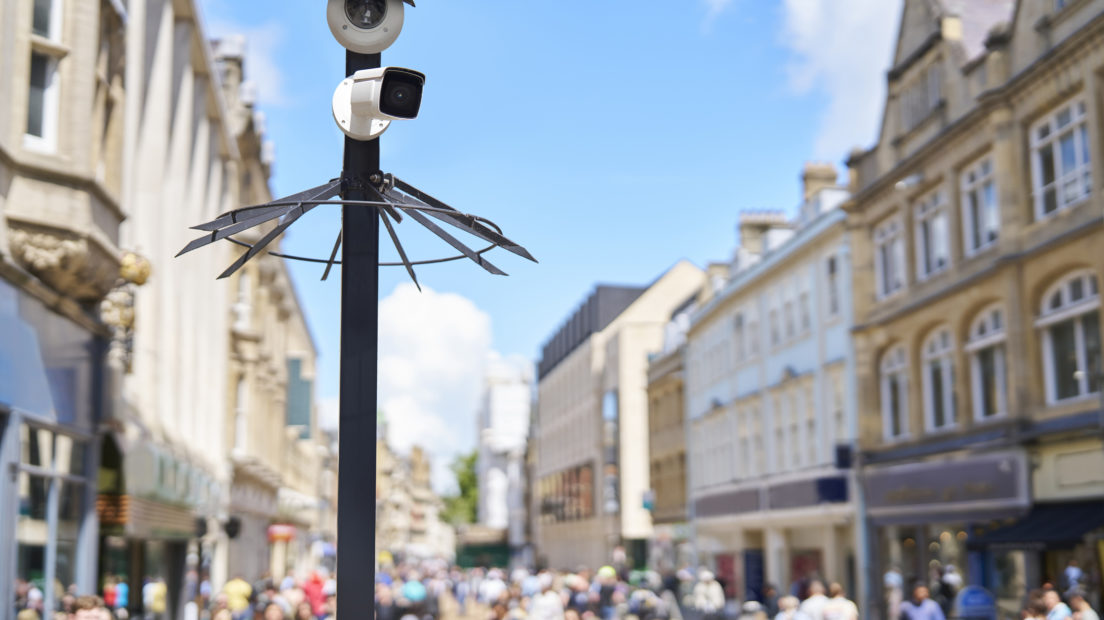 A CCTV security camera mounted on a post overlooking a busy high street